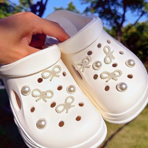 Può includere: Zoccoli bianchi con decorazioni a fiocco e perle decorative. Gli zoccoli hanno fori di ventilazione multipli e una suola spessa. I fiocchi e le perle sono disposti sulla parte superiore della scarpa. Gli zoccoli sono mostrati all'aperto.