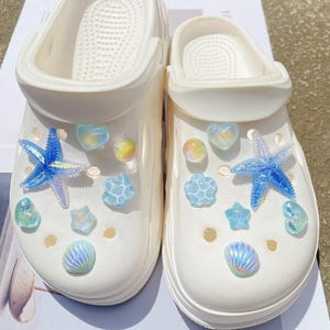 May include: White clogs decorated with blue starfish, seashell, and heart-shaped charms. The charms are iridescent and add a touch of sparkle. These beach-themed clogs are perfect for summer.