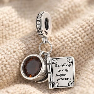 Può includere: Un charm in argento con una tazza di caffè, un libro e la scritta "Reading is my super power!". Il charm ha una parte superiore rotonda con la scritta "S925". La tazza di caffè ha una gemma marrone. Il libro ha quattro piccole gemme.