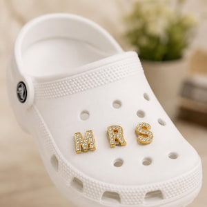 Può includere: Zoccolo bianco con lettere dorate di strass che compongono la parola "MRS". Le lettere sono attaccate alla parte superiore della scarpa. La scarpa ha una superficie strutturata e diversi fori di ventilazione.