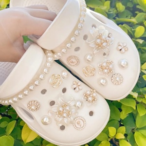 May include: White clogs decorated with pearl and rhinestone embellishments. The clogs have a pearl trim and decorative floral and circular accents. The shoes are set against a backdrop of green foliage.