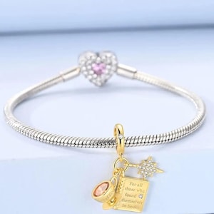 以下が含まれることがあります： ハート型のチャームと、ティーカップ、本、星型のチャームが付いたシルバーのチャームブレスレット。本のチャームには「For all those who found themselves in books」と書かれています。