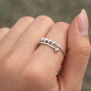 May include: A silver-colored ring with a unique design. The ring features a curved band with five small, round beads. The ring is worn on a finger, showcasing its minimalist and modern aesthetic. The ring is made of a shiny metal.