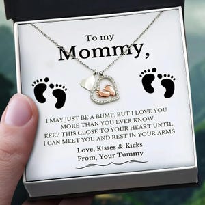 Op de afbeelding: Een zilverkleurige ketting met een hartvormige hanger met kleine babyvoetjes en heldere steentjes, gepresenteerd in een doosje met de tekst: To my Mommy, I may just be a bump, but I love you more than you ever know. Love, Kisses & Kicks From, Your Tummy.