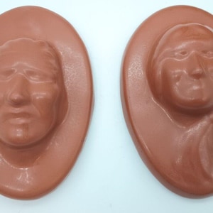 Op de afbeelding: Twee ovale, terracotta-kleurige plaquettes, elk voorzien van een verhoogd reliëfportret van een menselijk gezicht. De linker plaquette toont een gezicht met kort haar, terwijl de rechter plaquette een gezicht met lang haar weergeeft.