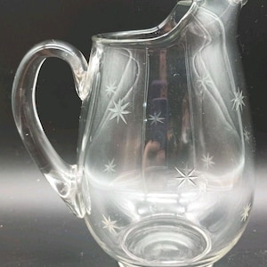 May include: Clear glass pitcher with a rounded body, a curved handle, and a pouring spout. The pitcher is decorated with etched starburst designs. The pitcher is designed for serving beverages.