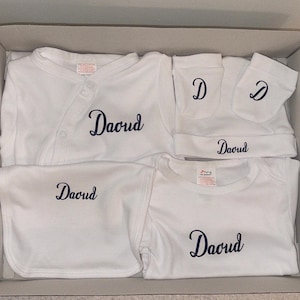 May include: A white baby clothing set presented in a cardboard box. The set includes a long-sleeved top, bib, short-sleeved top, hat, and mittens. Each item is embroidered with the name "David" in a dark blue script.