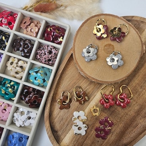 Pode incluir: Uma coleção de brincos em forma de flor em várias cores, incluindo vermelho, branco, castanho e prateado, exibidos numa bandeja de madeira e numa caixa dividida. Alguns brincos têm aros ou detalhes dourados. Os brincos são uma mistura de cores sólidas e glitter.