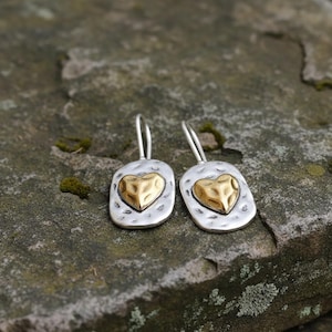 Puede incluir: Un par de pendientes de plata con un diseño de corazón dorado. Los pendientes tienen una textura martillada y una forma rectangular. El corazón dorado está centrado en el fondo plateado. Los pendientes se muestran sobre una superficie de piedra.