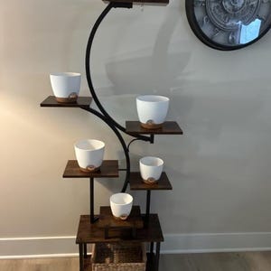 以下が含まれることがあります： 黒い金属フレームと濃い茶色の木製棚を備えた、多層のプラントスタンド。明るい茶色のベースが付いた白いセラミックポットが棚に置かれています。織りのバスケットがスタンドのベースにあります。装飾的な時計が壁に掛かっています。