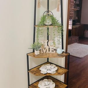 Peut inclure: Étagère d'angle avec une structure en métal noir et des étagères en bois. Les étagères sont décorées de bougies, de verdure et d'objets décoratifs, dont le mot "HOME" et des fleurs en papier. L'unité est placée dans un coin de la pièce.