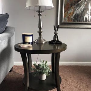 Może przedstawiać: Ciemnobrązowy, okrągły stolik boczny z dolną półką. Biały abażur spoczywa na srebrnej lampie. Mała czarna statuetka królika i niebieska świeca znajdują się na stole. Mała roślina w białej doniczce stoi na dolnej półce.