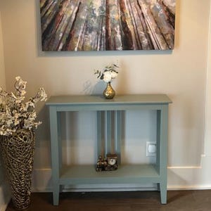 May include: A light blue console table with a lower shelf, holding a decorative clock and a gold vase with white flowers. A large gold vase with white flowers is on the left. A painting of a forest hangs above the table.