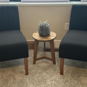 Puede incluir: Una pequeña mesa auxiliar redonda de madera con tres patas, colocada entre dos sillas negras tapizadas. Una maceta decorativa gris con vegetación se encuentra sobre la mesa. Las sillas y la mesa están sobre una alfombra de color neutro.