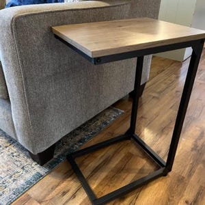 May include: A side table with a light brown tabletop and a black metal frame. The table is designed to slide under a sofa or chair. The tabletop is square and the frame has a C-shape design.