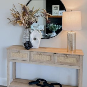 Puede incluir: Una consola de madera clara con tres cajones e inserciones de caña tejida. Un espejo negro circular refleja una habitación con chimenea. Un jarrón de flores secas, una escultura blanca y una lámpara con base dorada están sobre la mesa.