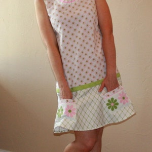 Puede incluir: Una mujer con un vestido blanco con detalles florales rosas y verdes y sandalias blancas. El vestido tiene un estampado de cuadros verdes y blancos en la mitad inferior.