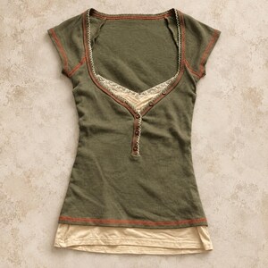 Pode incluir: Uma blusa verde-oliva de manga curta com decote redondo e acabamento em renda creme, uma carcela parcial com três pequenos botões e costuras expostas na cor ferrugem ao longo das costuras, sobreposta a uma bainha lisa na cor creme.