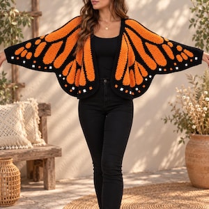Pode incluir: Um cardigã de crochê com um padrão de asas de borboleta monarca em laranja e preto. A peça tem um design aberto na frente com mangas largas em forma de asas e pequenos detalhes quadrados brancos ao longo das bordas pretas das asas.