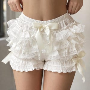 Puede incluir: Pantalones cortos blancos con volantes, ribete de encaje y lazos de satén. Los pantalones cortos presentan múltiples capas de tela, creando un efecto voluminoso. Los lazos están atados a la cintura y a los lados. Hechos de material suave y ligero.