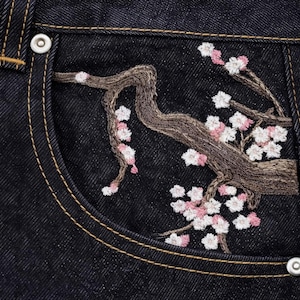 May include: Close-up of dark wash denim jeans pocket with embroidered cherry blossom branch. The embroidery features a brown branch with pink and white flowers. The pocket has a yellow stitch and a silver button.