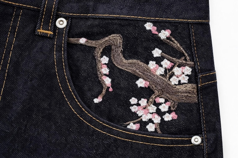 May include: Close-up of dark wash denim jeans pocket with embroidered cherry blossom branch. The embroidery features a brown branch with pink and white flowers. The pocket has a yellow stitch and a silver button.