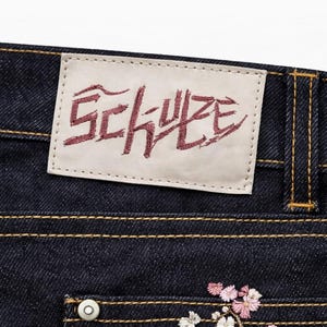 May include: Close-up of dark blue denim jeans with a leather patch featuring the word "SCHULZE" in burgundy lettering. The jeans have gold stitching and a small floral embroidery near the pocket.