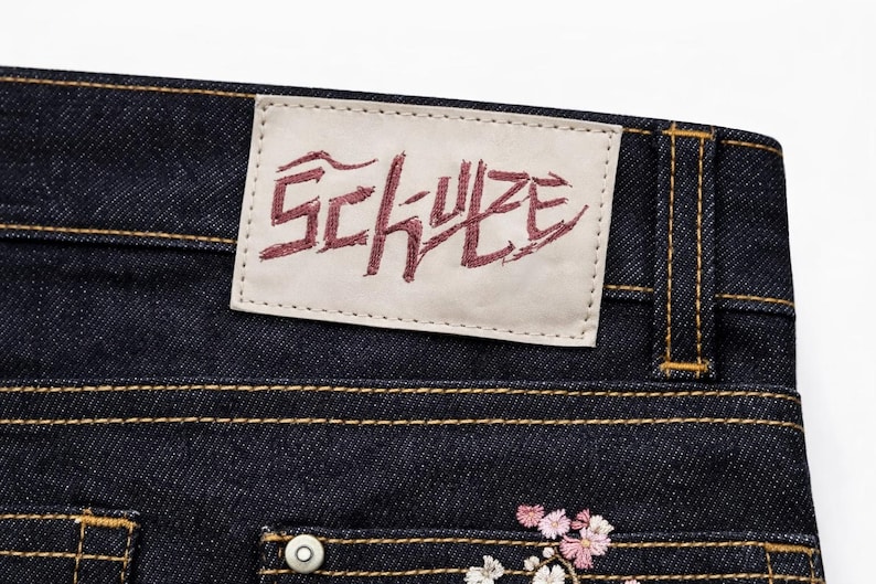 May include: Close-up of dark blue denim jeans with a leather patch featuring the word "SCHULZE" in burgundy lettering. The jeans have gold stitching and a small floral embroidery near the pocket.