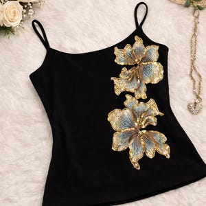 May include: Black camisole top with thin straps, adorned with two large, sequined floral designs in gold and blue. The top is made of a soft, dark fabric, perfect for a night out or special occasion.