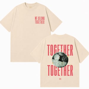 May include: Cream-colored t-shirt with short sleeves. The front features the text "WE BELONG TOGETHER" in red. The back displays the words "TOGETHER" twice, a disco ball graphic, and the year "2023" in red.
