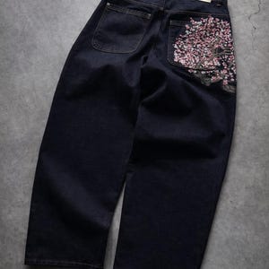 May include: Dark wash denim jeans with a wide leg cut. The back right pocket features an embroidered cherry blossom design in pink and white. The jeans have two back pockets and a label that reads "5252 by o!oi".