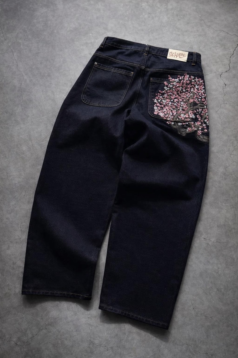 May include: Dark wash denim jeans with a wide leg cut. The back right pocket features an embroidered cherry blossom design in pink and white. The jeans have two back pockets and a label that reads "5252 by o!oi".