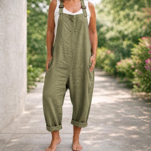 Könnte beinhalten: Olivgrüner Leinen-Jumpsuit mit verstellbaren Trägern und großen Vordertaschen. Das Design mit verkürztem, weitem Bein hat umgeschlagene Manschetten. Das Model trägt ein weißes Tanktop darunter. Der Jumpsuit hat eine lockere Passform.