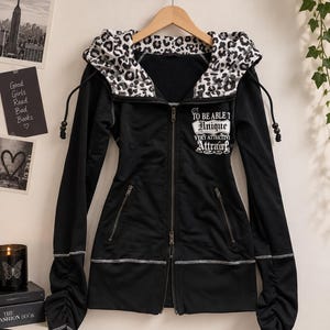 May include: A black zip-up hoodie featuring a white leopard print lining on the hood, long sleeves with ruched detailing, and two zippered pockets. The chest displays white text reading TO BE ABLE TO Unique VERY ATTRACTIVE Attractive.