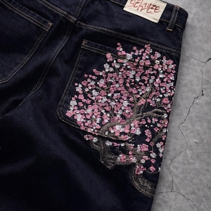 May include: Dark wash denim jeans with a floral embroidery design on the back pocket. The embroidery features a tree with pink and white blossoms. A rectangular label with text is visible on the back.