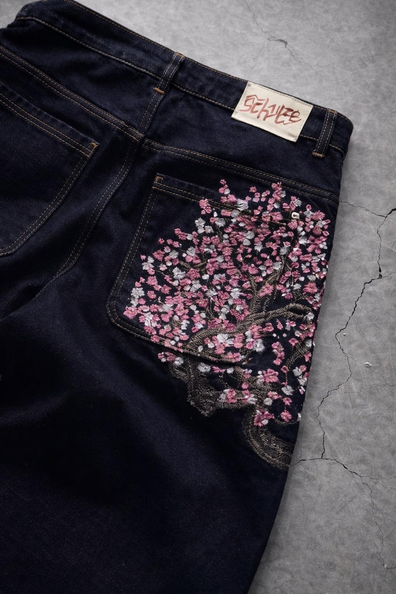May include: Dark wash denim jeans with a floral embroidery design on the back pocket. The embroidery features a tree with pink and white blossoms. A rectangular label with text is visible on the back.