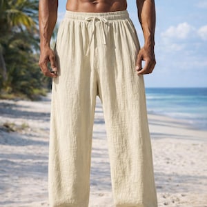 May include: Beige linen pants with a drawstring waist. The pants are loose-fitting and have a relaxed style. The model is standing on a sandy beach with the ocean in the background. The pants are a neutral color, suitable for casual wear.