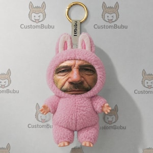 Könnte beinhalten: Ein Plüsch-Schlüsselanhänger in Form eines flauschigen rosa Hasenkostüms mit einem realistischen menschlichen Gesicht in der Mitte. Das Spielzeug hat kleine Hände und Füße aus Kunststoff und ist mit einem grauen Stoffband mit CustomBubu-Aufdruck befestigt.