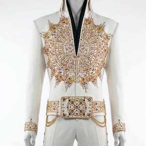 Könnte beinhalten: Weißer Overall mit aufwendigen Gold- und Juwelenverzierungen. Die Jacke hat einen hohen Kragen und ein zentrales Design aus konzentrischen Kreisen und strahlenförmigen Mustern. Der passende Gürtel ist ebenfalls stark verziert, präsentiert auf einer Schaufensterpuppe.