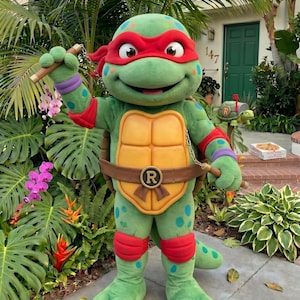 Può includere: Una persona in costume da Raffaello delle Tartarughe Ninja. Il costume è verde con una maschera rossa, ginocchiere e bracciali. Il personaggio tiene in mano dei nunchaku e ha una cintura marrone con una fibbia dorata. Lo sfondo comprende una vegetazione lussureggiante e una casa.