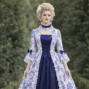 Puede incluir: Una mujer con un vestido estampado floral azul y blanco. El vestido presenta un corpiño azul marino con cordones en la parte delantera y una falda a juego. La capa exterior es blanca con diseños florales azules y bordes con volantes. Las mangas tienen lazos azules.