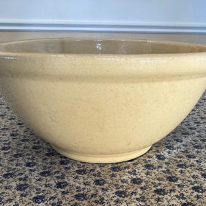 May include: A large, cream-colored stoneware bowl with a wide, rounded shape. The bowl has a smooth, slightly speckled surface and a thick rim. It sits on a patterned tablecloth, suggesting a kitchen or dining setting. This bowl is suitable for mixing or serving.