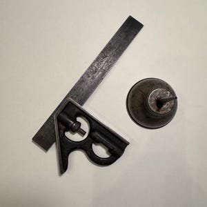 May include: A vintage metal combination square with a dark, weathered finish and visible inch markings on the blade, positioned next to a small, round, dark grey metal oil can with a pointed spout and a textured, aged surface appearance.