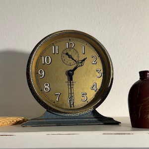 May include: An antique gold-toned clock with black hands and numerals, set on a blue base. A small brown jug with a handle and decorative gold accents sits to the right. The clock face reads "Westclox".