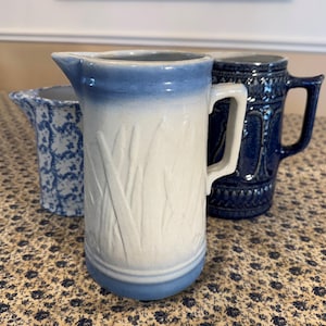 May include: Three ceramic pitchers in varying shades of blue and white. The central pitcher is tall, with a white body and blue rim, featuring an embossed wheat design. A smaller pitcher with a blue floral pattern and a dark blue mug are also present.