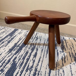 May include: A small, round, brown wooden stool featuring three cylindrical legs and a single, horizontal handle extending from the side of the seat. The stool has a smooth, polished finish and is constructed entirely from a solid, medium-toned wood.