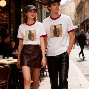 May include: A couple wearing matching white t-shirts with red trim and a graphic print. The woman wears a brown leather skirt, and the man wears black pants. Both wear caps and walk down a city street.