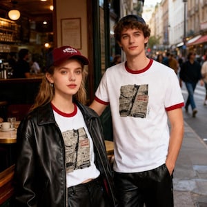 May include: A couple wearing matching white t-shirts with a red trim and a graphic print. The woman wears a black leather jacket and shorts, and a burgundy baseball cap. The man wears black pants. They are standing outside a cafe.