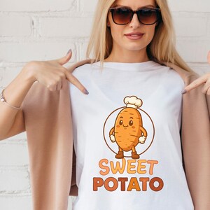 May include: White V-neck t-shirt featuring a cartoon sweet potato character wearing a chef's hat, inside a brown circle. The words "SWEET POTATO" are printed in orange and brown below the graphic.