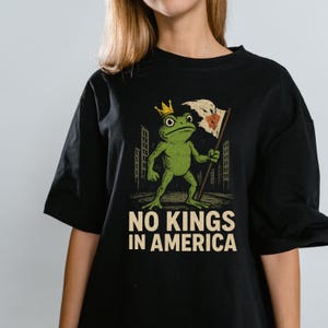 Puede incluir: Camiseta negra con una ilustración de una rana verde con una corona dorada y una bandera con un diseño de corazón. El texto "NO KINGS IN AMERICA" está impreso debajo de la ilustración de la rana. La camiseta tiene un corte holgado.